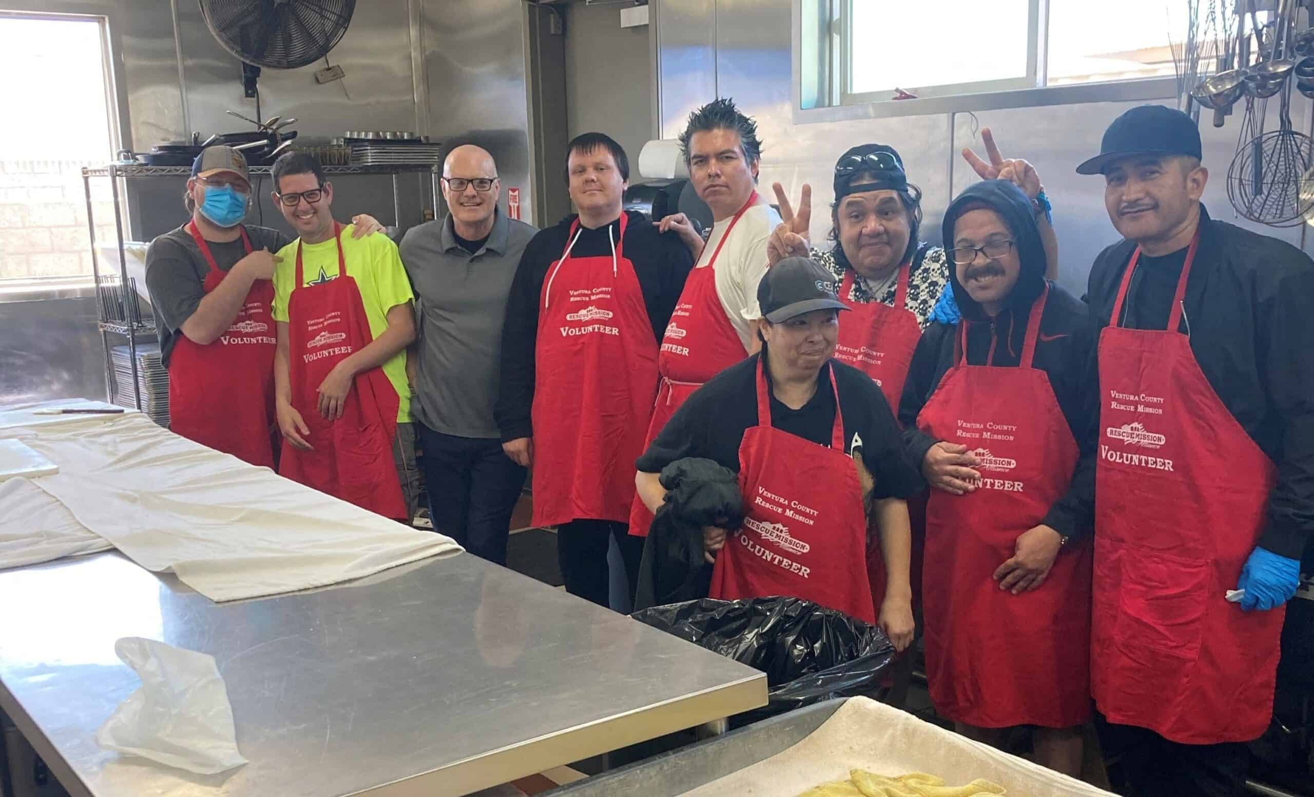 A group of people wearing red volunteer aprons pose together in a commercial kitchen, standing behind a metal counter with baking trays.