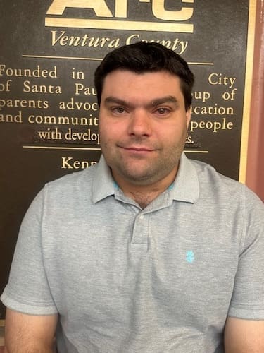 A man in a light gray polo shirt sits in front of a plaque that has text about Ventura County.