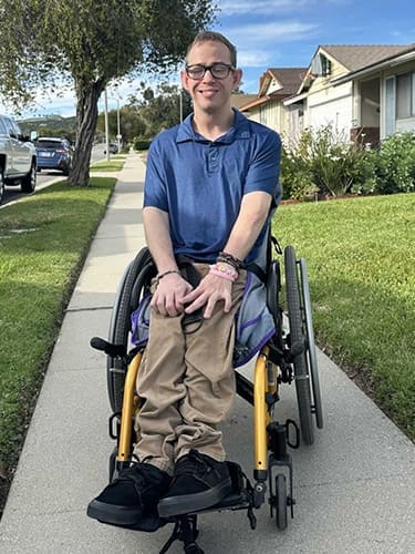 A person in a wheelchair sits on a sidewalk, wearing glasses, a blue shirt, and khaki pants, smiling at the camera. Houses and grass are visible in the background.