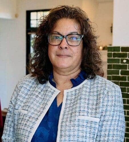 A woman with curly brown hair and glasses wearing a blue blouse and a light plaid jacket stands indoors, facing the camera.