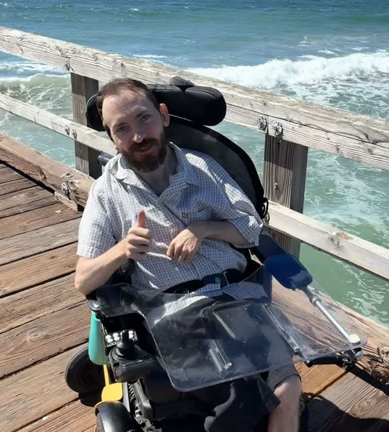 Man in a wheelchair gives a thumbs-up while sitting on a wooden pier by the ocean on a sunny day.
